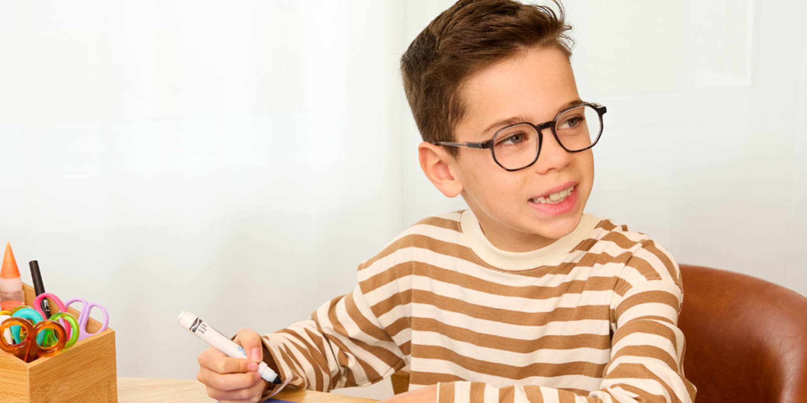 male child with glasses smiling while colouring wearing a orange striped long sleeve shirt