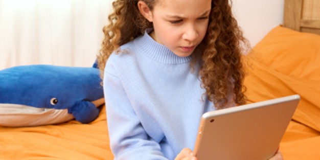 young girl sitting on a bedf and squinting while looking at an Ipad